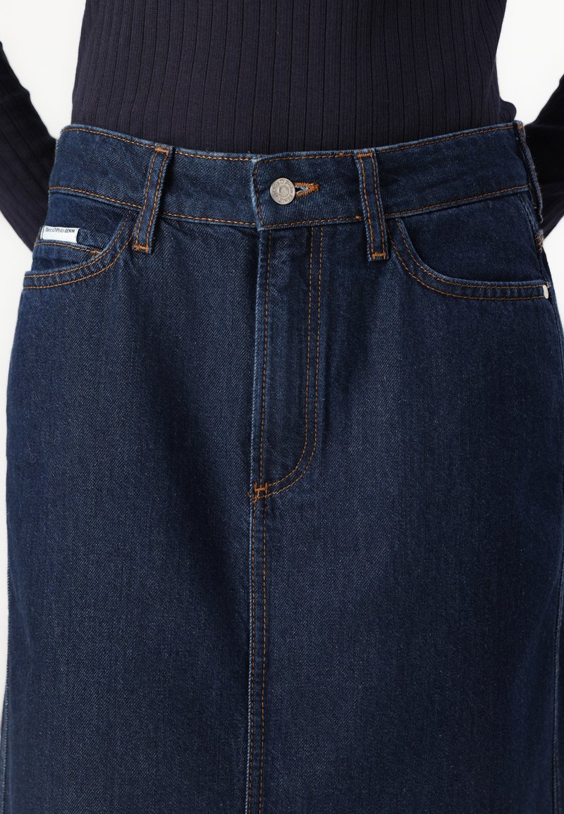 Denimrock mit einer dunklen Indigo-Waschung, sichtbaren orangenen Nähten, vorderen Taschen und einem Frontknopfverschluss. Glatte Oberfläche.