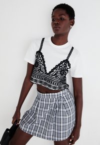 Camiseta blanca con superposición de encaje negro recortada, que presenta intrincados patrones florales. Combinada con una falda corta de cuadros plisada en gris y negro.