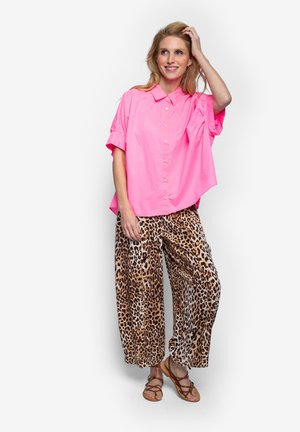 Vrouw in een felroze oversized blouse, wijde broek met luipaardprint en bruine sandalen met bandjes, glimlachend terwijl ze met haar hand haar haar aanraakt.