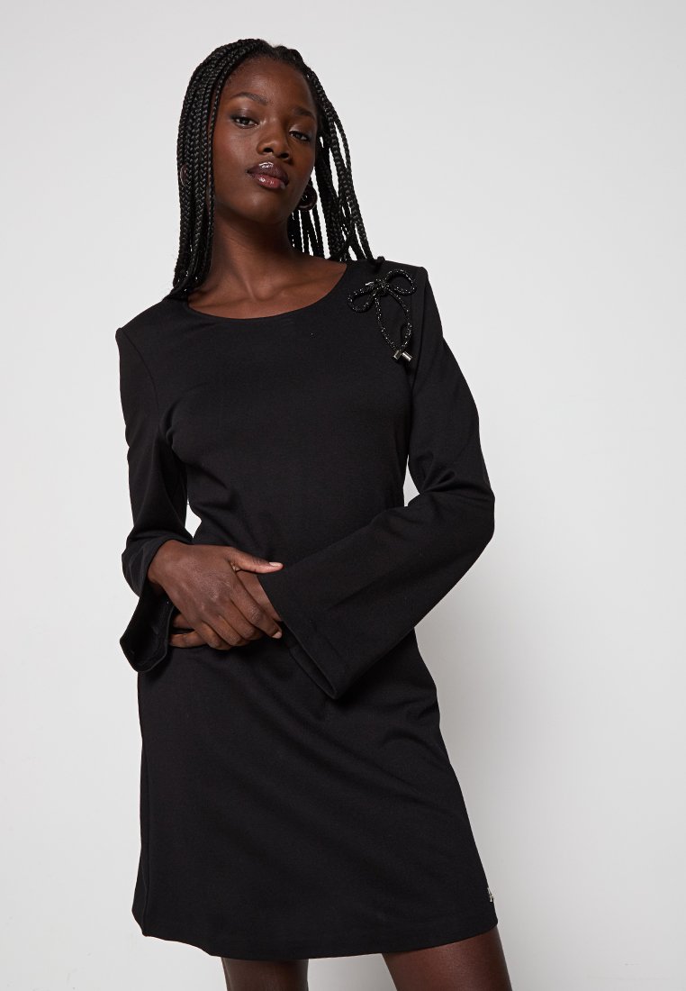 Kobieta z warkoczami, ubrana w czarną sukienkę z długim rękawem i rozkloszowanymi mankietami oraz ozdobnym detalem w postaci kokardy na ramieniu, pozująca na jednolitym tle.