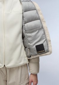 Lettet jakke med en cremefarvet fleece-krop, grå pufferærme, teksturerede detaljer og et sort mærke med logo. Lukkemekanismen har en lynlås.