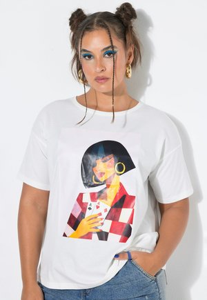 Femme aux cheveux tressés en chignons porte un t-shirt blanc avec un graphisme d'une femme tenant des cartes à jouer, associé à un jean bleu.