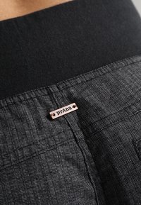 Gros plan d'un tissu gris foncé texturé avec une petite étiquette métallique rectangulaire portant la marque "prAna" cousue près d'une couture.