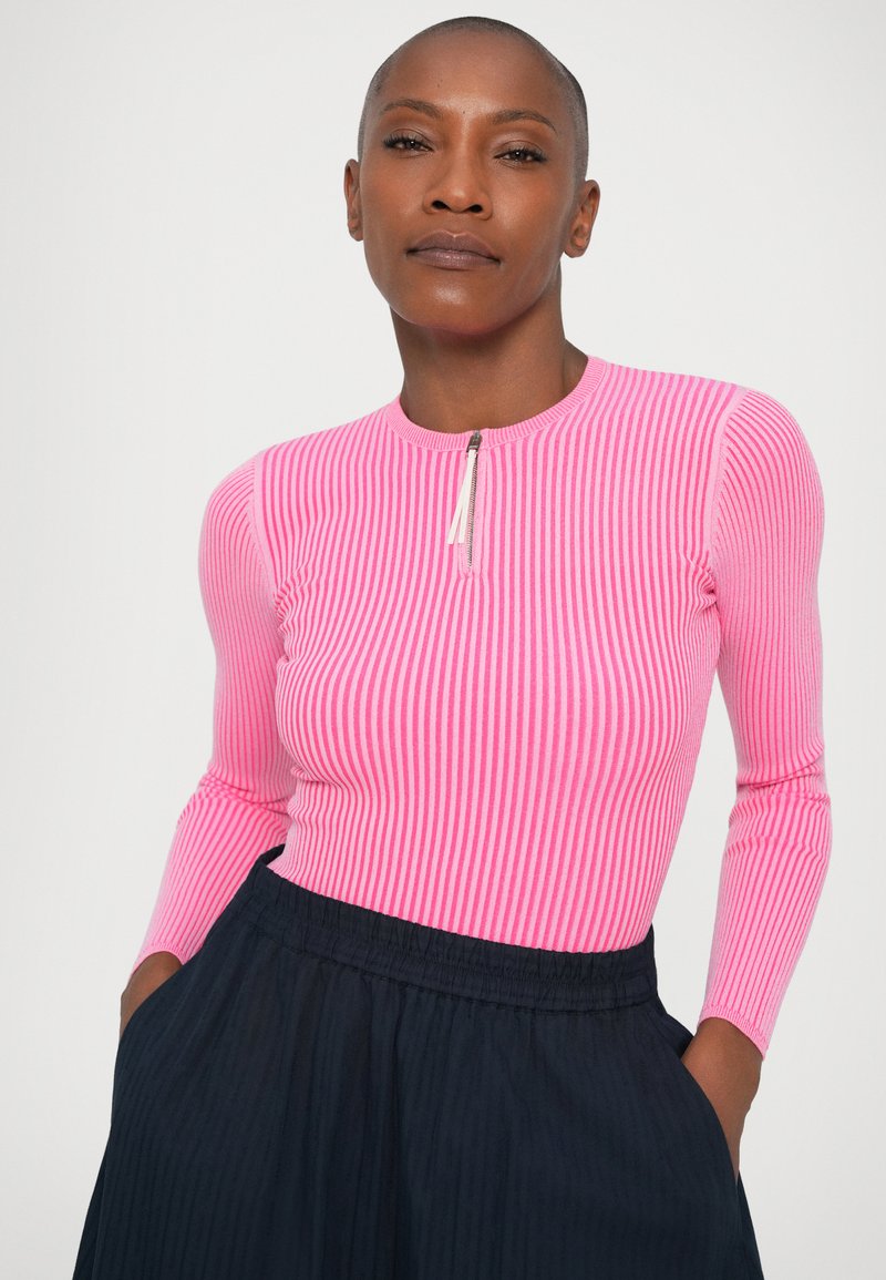Ribbed long-sleeve top in bright pink, featuring a round neckline and a short front zipper. Paired with a dark blue skirt.