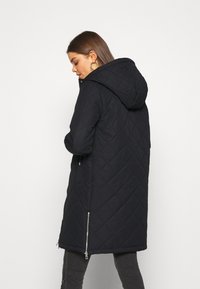 Manteau noir matelassé avec une capuche, présentant une fermeture éclair et un motif de couture en diagonale. Il a une coupe mi-longue et des accents de fermeture éclair en argent.