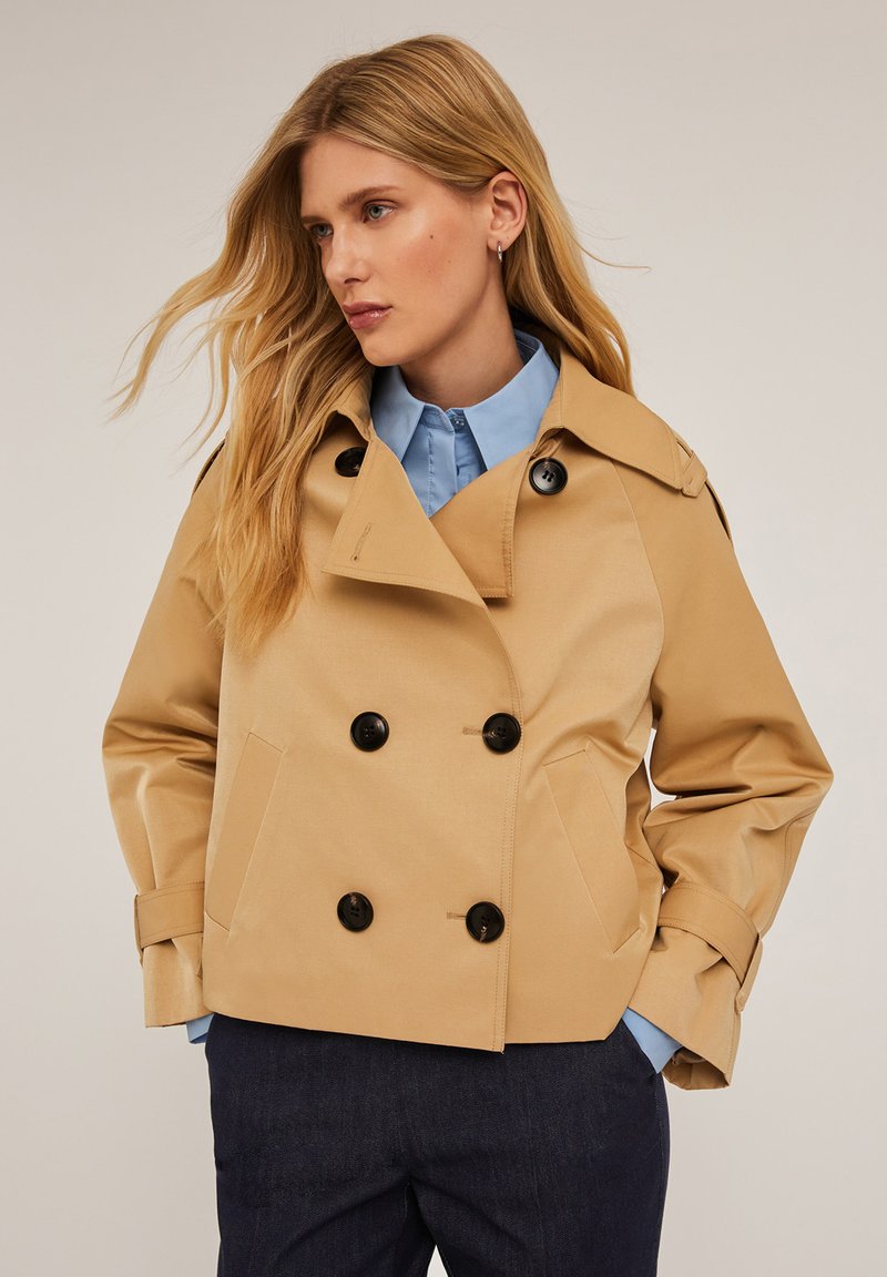Trench coat de color beige con doble botonadura y botones negros, que presenta un cuello estructurado y bolsillos laterales. Lleva puesto una camisa de color azul claro.