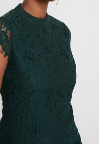Tmavě zelené krajkové šaty s vysokým výstřihem a krátkými rukávy, které nosí osoba s hnědou pletí a zlatými visacími náušnicemi, na bílém pozadí.