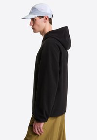 Μαύρο φλις φούτερ με φαρδιά εφαρμογή και κουκούλα, φορεμένο με ανοιχτό μπλε καπέλο από δίχτυ και μπεζ παντελόνι, που αναδεικνύει την υφή και τον καθημερινό σχεδιασμό.