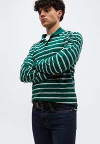 Νεαρός με σγουρά μαλλιά που φοράει μακρυμάνικη πόλο μπλούζα με πράσινες και λευκές ρίγες και σκούρο τζιν, με σταυρωμένα χέρια, κοιτάζοντας στο πλάι.