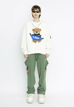 Sudadera blanca con un gráfico de un oso de peluche y una bolsa azul. Pantalones cargo verde oliva con bolsillos. Zapatillas blancas gruesas. Gafas de sol negras.