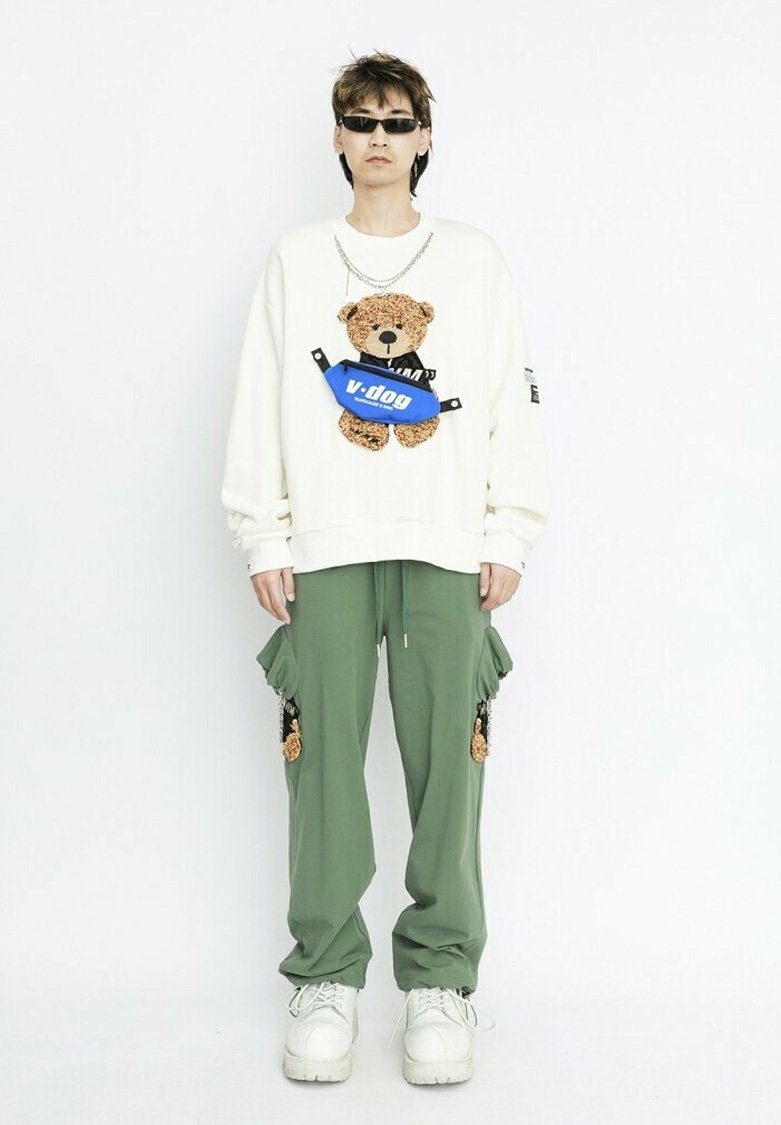 Sudadera blanca con un gráfico de un oso de peluche y una bolsa azul. Pantalones cargo verde oliva con bolsillos. Zapatillas blancas gruesas. Gafas de sol negras.