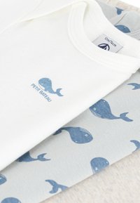 Hvit bomullsheldress med grafikk av en blåhval og teksten "PETIT BATEAU", sammen med et lys grått stoff som viser et hvalmønster.