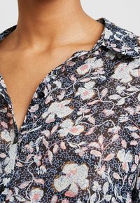 Blommig skjorta med knappar i genomskinlig svart tyg. Har ett flerfärgat blommönster i blå, rosa och vita nyanser. Klassisk kragdesign.