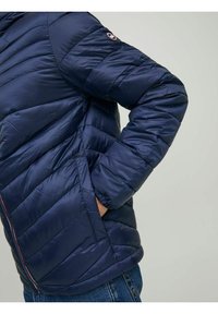 Tmavě modrá prošívaná bunda s kapucí, která má zip a boční kapsy. Vyrobena z lesklého, hladkého materiálu s vyšívaným logem.