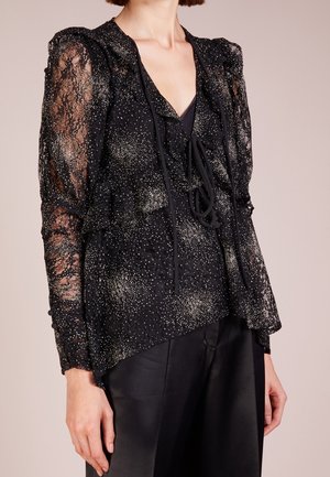 Schwarze transparente Spitzenbluse mit metallischen Sprenkeln, Bindekragen, geraffter Vorderseite und langen Ärmeln, kombiniert mit schwarzen Hosen.