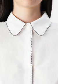 Primer plano de una camisa blanca con un collar con ondas y ribete negro, que presenta botones en la parte delantera, destacando sus delicados detalles.