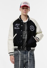 Veste varsity noire et crème avec texte brodé, boutons-pression, poignets rayés, portée sur un t-shirt gris et associée à un jean déchiré.