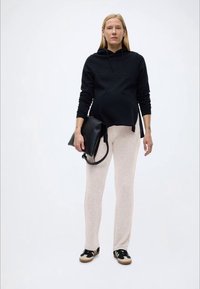 Έγκυος γυναίκα όρθια, φοράει μαύρο φούτερ με κουκούλα, μπεζ παντελόνια, μαύρα και λευκά αθλητικά παπούτσια, και κρατάει μια μαύρη δερμάτινη τσάντα.