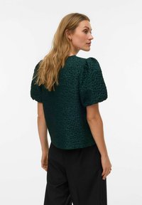 Rohelise tekstuuriga pluus lühikeste, volangidega varrukatega, ümara kaelusega ja lõdvestunud lõikega. Sobitatud mustade pükstega.