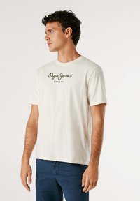 Homme portant un t-shirt crème Pepe Jeans London et un pantalon bleu marine, se tenant debout et regardant vers la gauche sur un fond blanc uni.