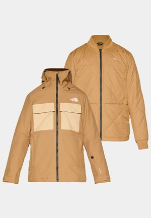 Bruine outdoorjas met een afneembare capuchon en twee grote voorzakken. De binnenkant heeft een geborduurd ontwerp in een vergelijkbare kleur.