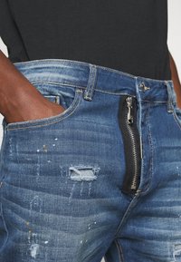 Vaqueros de mezclilla en azul con detalles desgastados, incluyendo desgarros y manchas de pintura. Presentan un zipper lateral negro y trabillas para cinturón estándar.