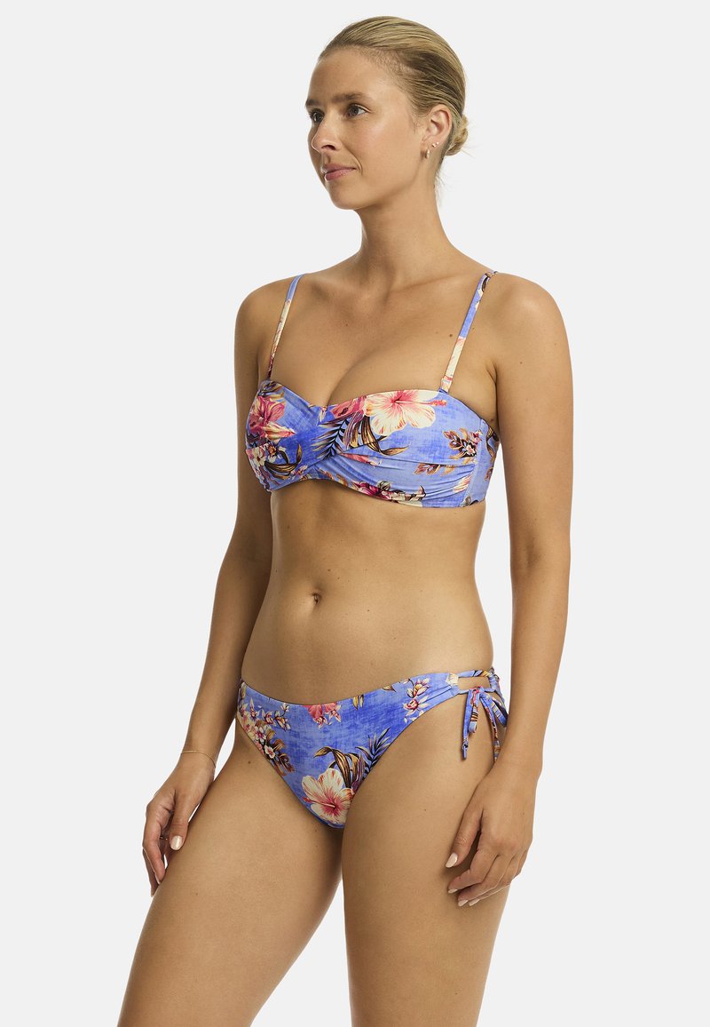 Eine Frau steht selbstbewusst da, trägt einen blauem Bikini mit floralem Muster und dünnen Trägern, der tropische Farben und ein stylisches Design zeigt.