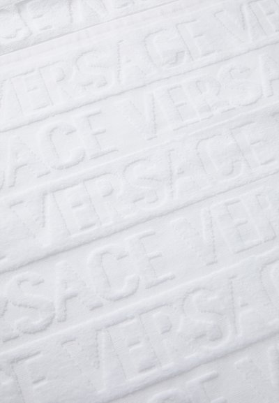 Serviette blanche ornée d'un motif en relief du mot "VERSACE" en lettres capitales, avec une finition douce et texturée.