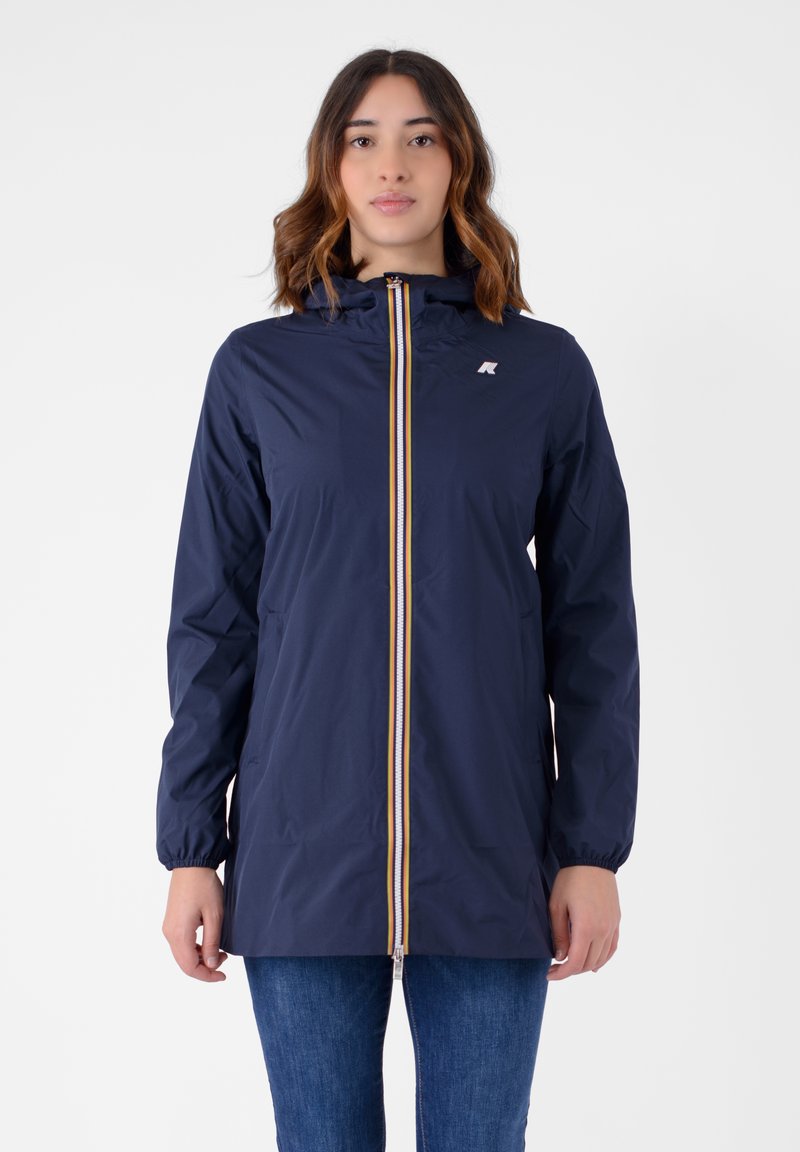 Giacca impermeabile blu navy con cappuccio, dotata di una cerniera gialla e bianca e polsini elasticizzati, indossata sopra un paio di jeans blu.