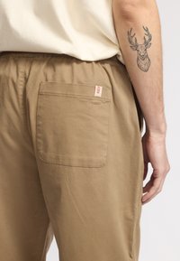 Beige katoenen broek met een elastische tailleband en een achterzak met een rood label. De stof heeft een gladde textuur.