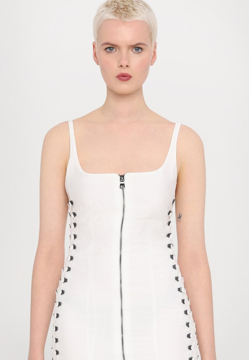 Witte aansluitende jurk met een vierkante halslijn, rits in het midden en zwarte veterdetails aan de zijkanten. Glad materiaal met minimale textuur.