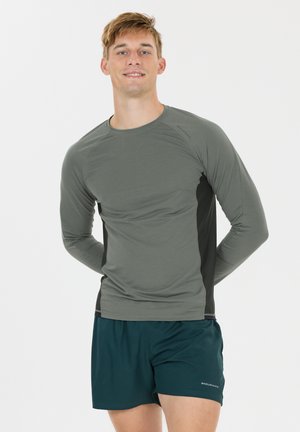 Jonge man lacht, draagt een grijs sportshirt met lange mouwen en donkergroene korte broek, staat met handen achter zijn rug tegen een witte achtergrond.