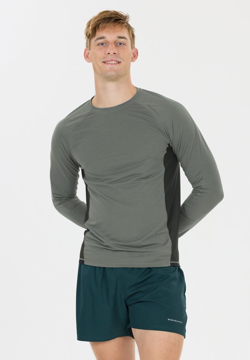 Ung mand smilende, iført grå langærmet træningsshirt og mørkegrønne shorts, stående med hænderne bag ryggen på hvid baggrund.