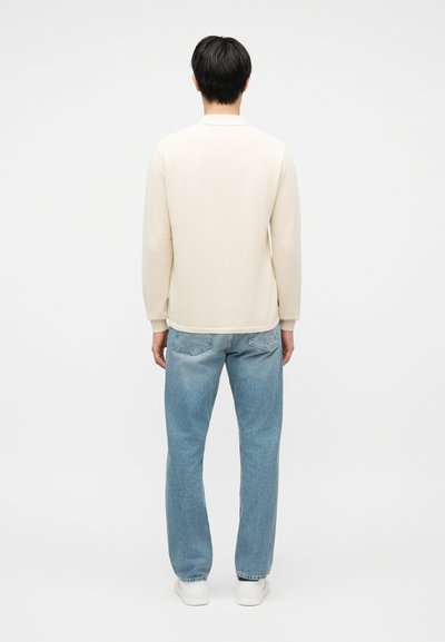 Homme debout de dos portant une chemise à manches longues crème, un jean bleu clair et des baskets blanches sur un fond blanc uni.