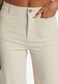 Beige high-waisted broek met een gladde textuur, voorzien van een ritssluiting aan de voorkant, gestikte zakken en een klein metalen logo-accent op de tailleband.