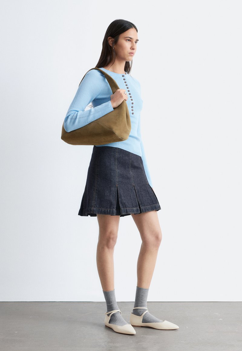 Femme portant un haut à manches longues bleu clair, jupe plissée foncée, chaussettes grises, ballerines crème, tenant un grand sac à épaule vert olive, debout de profil.