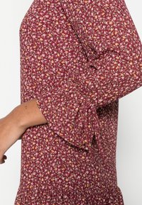 Ärmeldetail eines weinroten Kleides mit kleinen rosa und gelben Blumen, das einen ausgestellten Bund und gerafftes Stoffmaterial am Ellbogen aufweist.