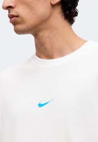Νέος άνδρας φοράει λευκό t-shirt με στρογγυλή λαιμόκοψη και μικρό, φθαρμένο μπλε λογότυπο Nike Swoosh στο στήθος.