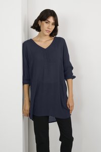 Tunic blu navy con scollo a V, maniche lunghe arrotolate, tessuto leggero e orlo asimmetrico, abbinata a pantaloni neri.