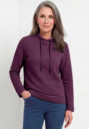 Vrouw van middelbare leeftijd met grijs haar draagt een paars gestructureerd capuchontrui en blauwe broek, staand met één hand in de zak tegen een witte achtergrond.