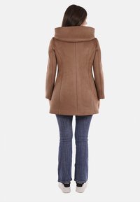 Cappotto peacoat in suede marrone con un ampio colletto ripiegato, silhouette aderente e dettagli delle cuciture. Abbinato a jeans blu e sneakers bianche.
