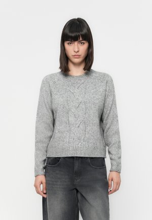 Mulher jovem com cabelo castanho escuro liso, vestindo um suéter cinza de malha trançada e jeans pretos desbotados, de pé contra um fundo branco simples.
