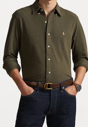 Man draagt een olijfgroen overhemd met knopen, donkerblauwe spijkerbroek, bruine leren riem en een polshorloge, staat met handen in de zakken.