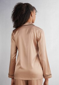 Långärmad satinpyjamasöverdel i beige, med en mjuk yta och kontrasterande svart pipning längs kragen och manschetterna. Bilden visar bakifrån.