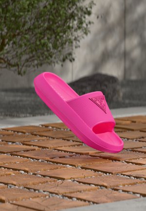 Șlapi roz aprins cu talpă texturată și logo-ul "Guess", așezați pe un suport din plăci de lemn, cu un fundal exterior estompat.