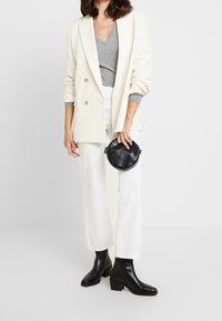 Crème dubbelknop blazer, grijze v-hals top, witte wijde broek, zwarte textuur cirkelvormige tas en zwarte enkelboots met een blokhak.