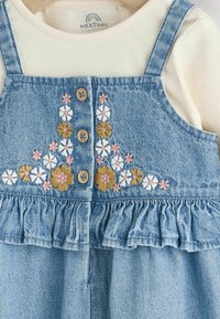 Vestido de mezclilla con bordados florales en rosa, amarillo y blanco, que presenta botones de madera y un dobladillo con volantes. Superpuesto sobre una blusa crema.