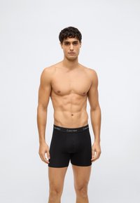 Man met kort krullend haar in zwarte Calvin Klein boxershorts, staand tegen een effen lichte achtergrond, met het gezicht naar voren.