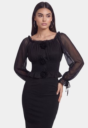 Donna con lunghi capelli scuri che indossa una camicetta nera trasparente a maniche lunghe con dettagli floreali e una gonna nera aderente, in piedi con una mano sul fianco.