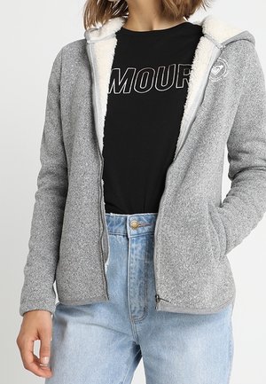 Femme portant une veste polaire gris clair zippée avec une doublure sherpa blanche sur un t-shirt noir avec le texte « AMOUR », assortie à un jean bleu clair.